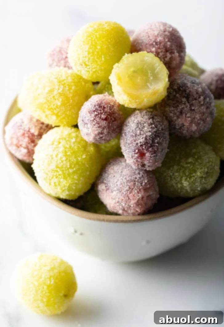 A vibrant bowl filled with both green and purple candied grapes. One green grape is shown bitten in half, revealing its juicy interior beneath the sparkling candy coating.