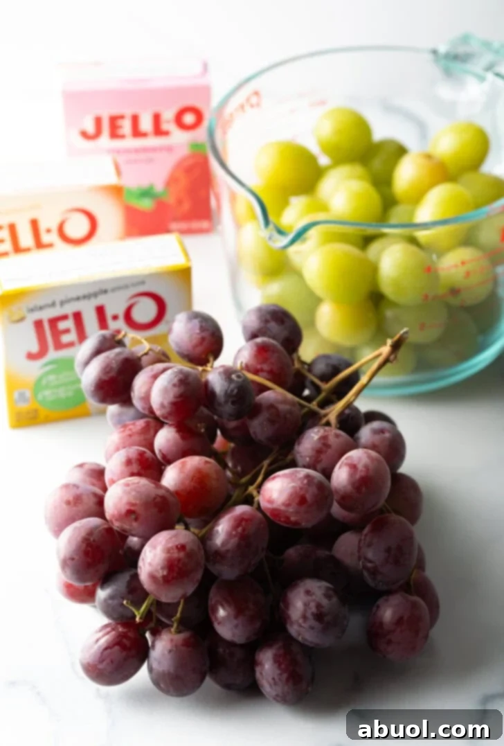 Recipe ingredients laid out: a vibrant bunch of red grapes prominently in the foreground, with a large glass measuring cup of fresh green grapes in the background, flanked by three colorful boxes of Jell-O.