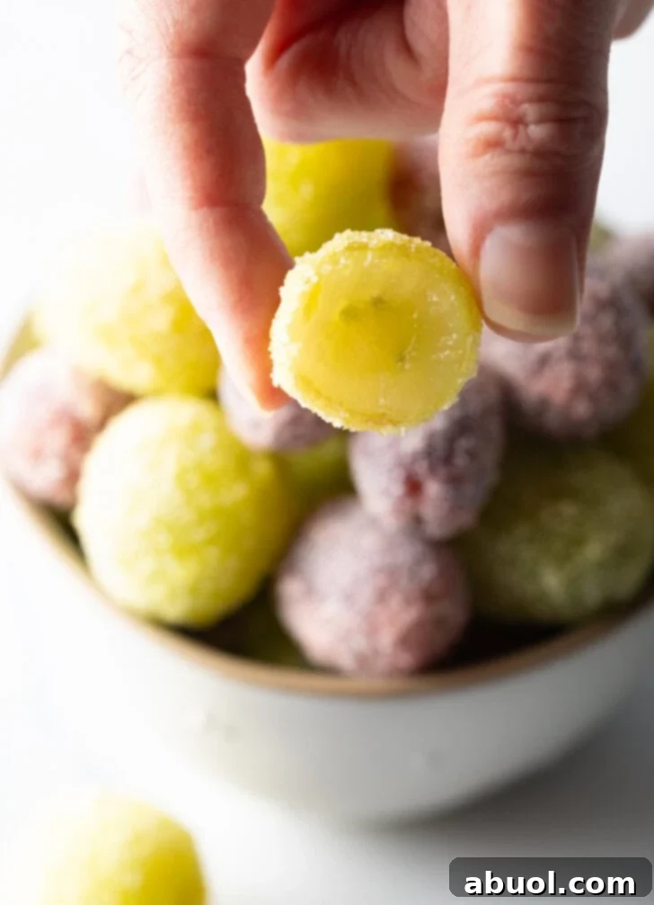 A close-up of a hand holding a green candy grape, which has been artfully bitten in half to reveal its fresh, juicy interior beneath the perfect candy shell, with a bowl of green and purple candy grapes blurred in the background.