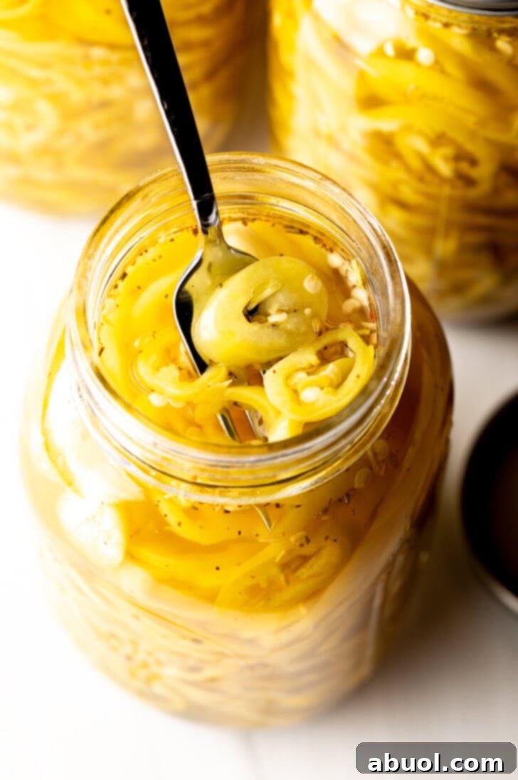 4 large jars of canned pickled banana peppers. The jar closest to the camera has the lid off showing the slices of peppers filling the jar. A fork is pulling a serving of slices from the jar.