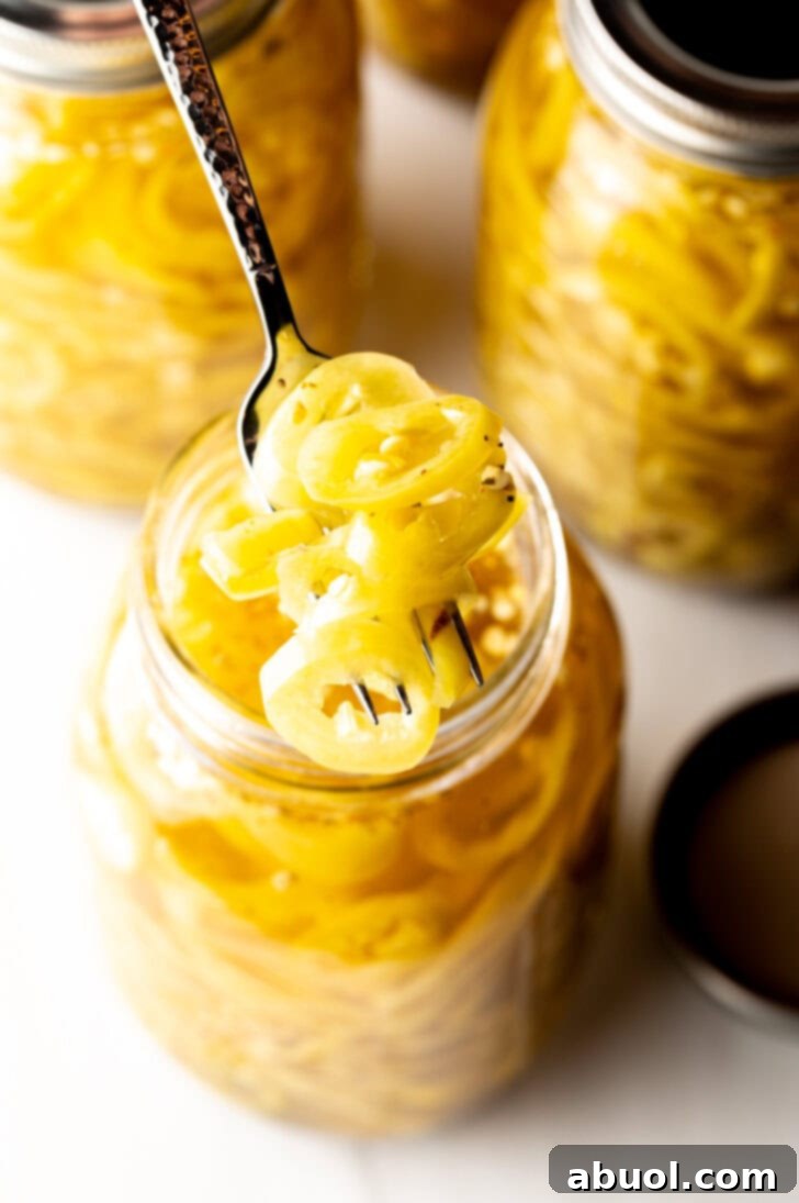4 large jars of canned pickled banana peppers. The jar closest to the camera has the lid off showing the slices of peppers filling the jar. A fork is pulling a serving of slices from the jar.