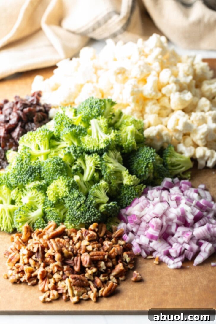 A cutting board neatly displaying individual piles of fresh broccoli florets, cauliflower florets, chopped red onions, toasted pecans, and plump dried cranberries, ready for assembly.