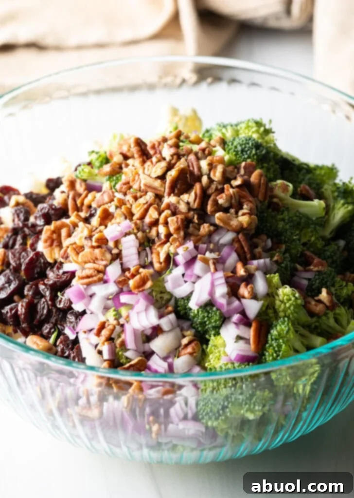 A clear glass bowl containing generous piles of vibrant fresh broccoli and cauliflower florets, alongside chopped red onions, pecans, and dried cranberries, prior to dressing.