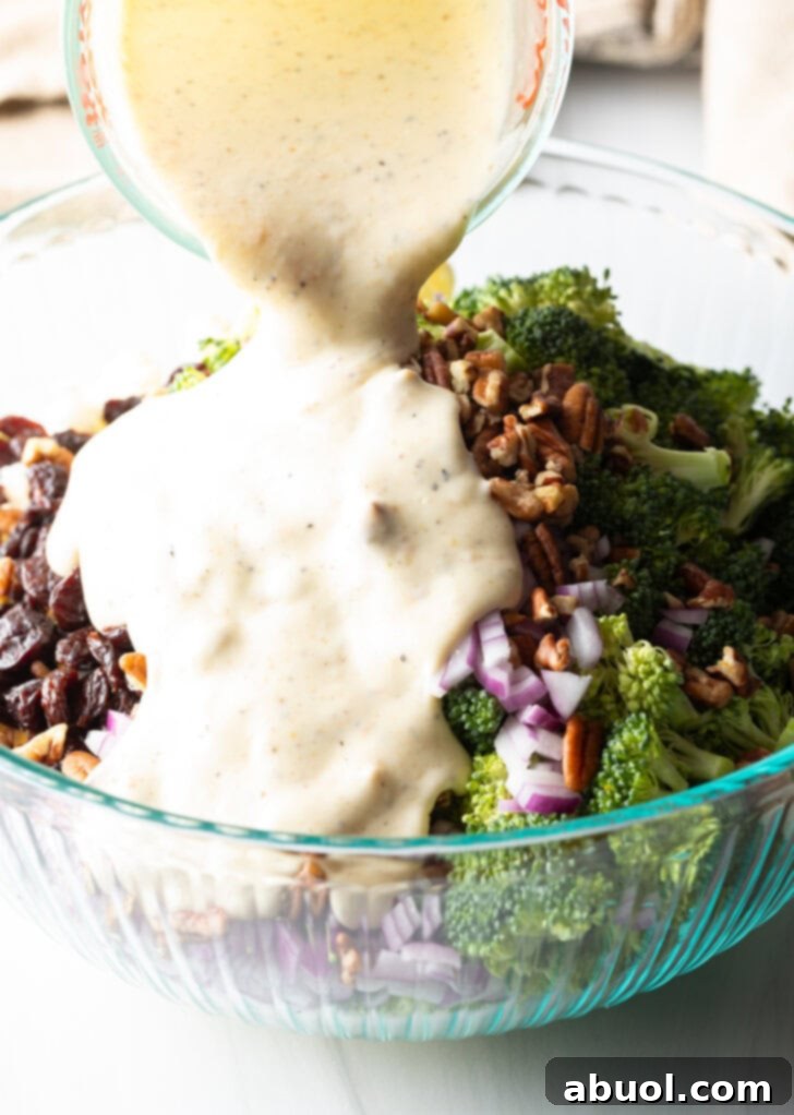 A smooth, creamy mayonnaise-based dressing being poured from a glass measuring cup directly onto the colorful mix of broccoli, cauliflower, nuts, and dried fruit in a large glass bowl.