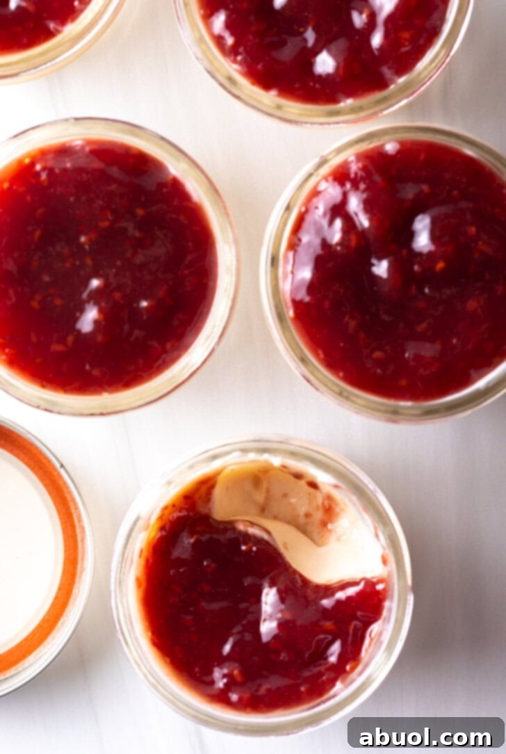 Overhead view of four elegant glass jars filled with creamy vanilla protein yogurt, each generously topped with vibrant red raspberry jam, showcasing a perfect healthy meal prep option or an appealing dessert.