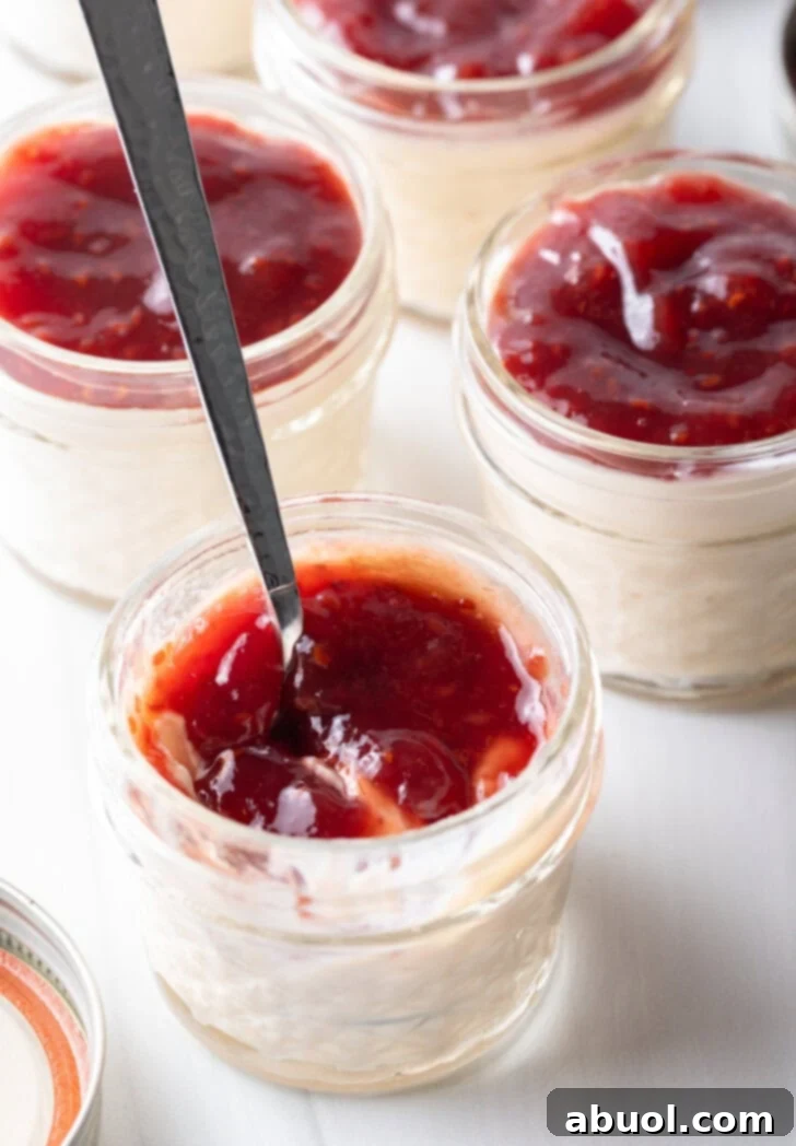 Top-down shot of four small glass jars of protein pudding, each perfectly topped with a dollop of vibrant raspberry jam. A metal spoon rests invitingly in the jar closest to the camera, hinting at the creamy texture and delicious flavor of this healthy treat.