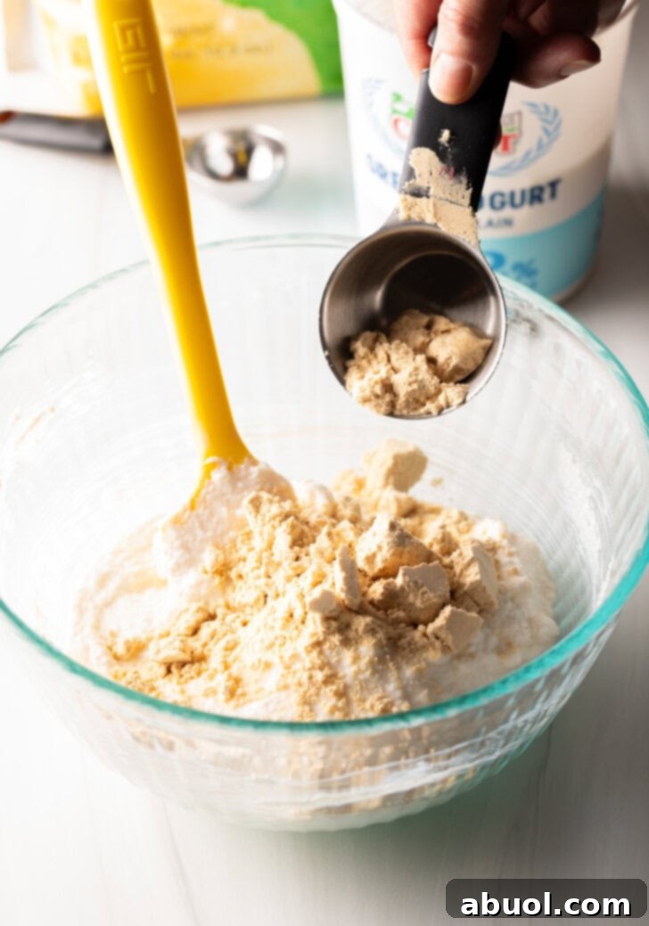 A metal measuring cup carefully pouring white vanilla protein powder into a large glass mixing bowl, where creamy Greek yogurt is already waiting to be combined for the protein pudding recipe, highlighting the active preparation process.