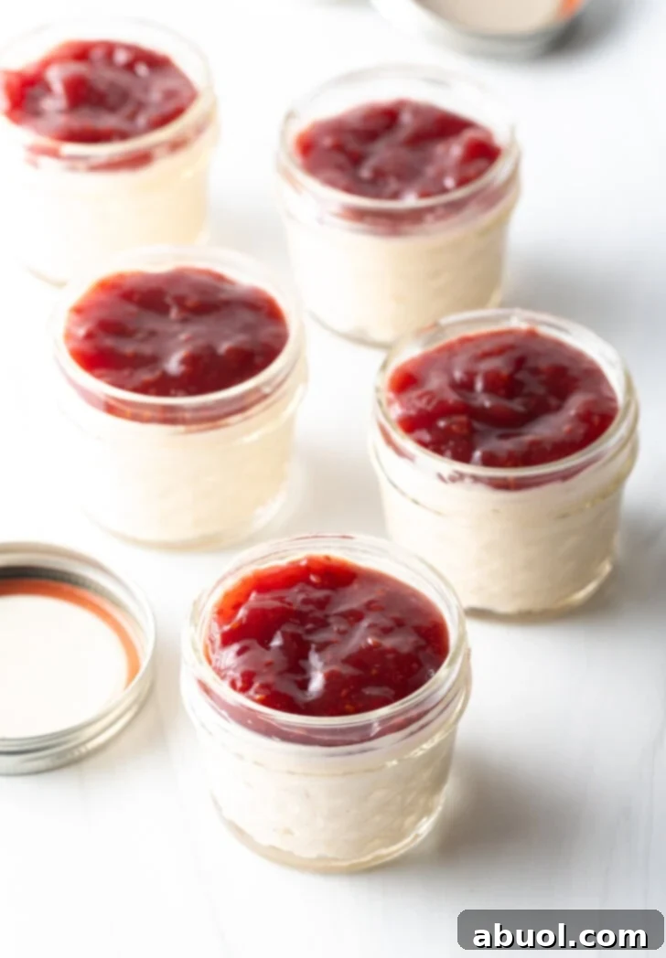 Five glass jars of protein pudding, each beautifully topped with vibrant raspberry jam, arranged on a rustic wooden surface. The composition highlights a healthy, visually appealing, and delicious dessert or snack option.