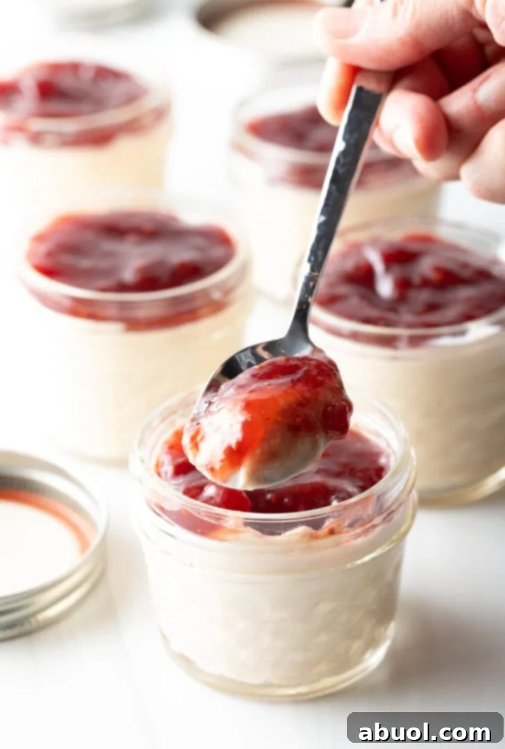 A metal spoon lifted from a glass jar of protein yogurt, showcasing a perfect scoop of creamy pudding elegantly mixed with bright red raspberry jam, inviting a delightful taste.