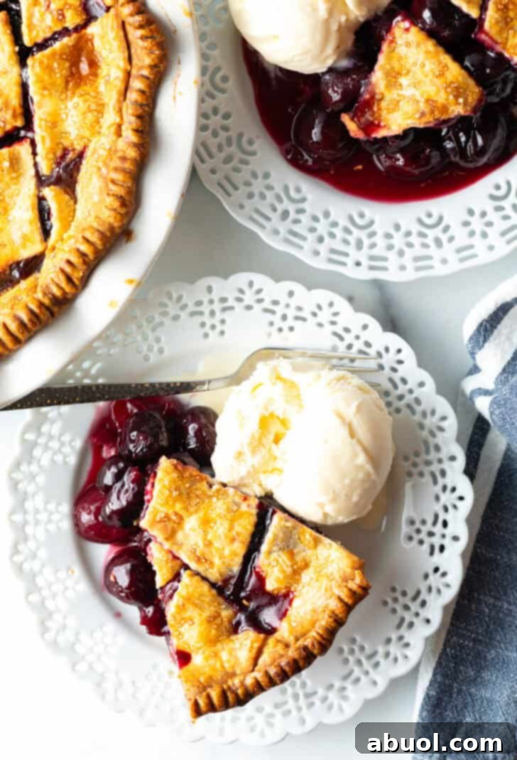 Top down view of a slice of homemade cherry pie, with cherry pie filling from scratch, on a white plate, with scoop of vanilla ice cream on the side.