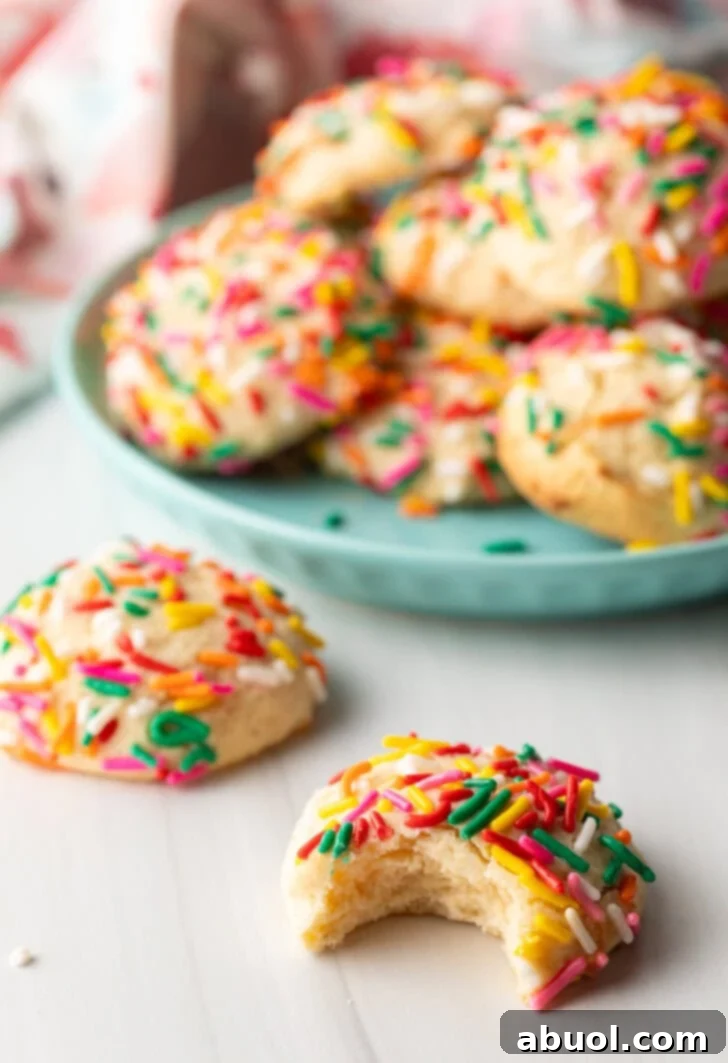 Luscious Cream Cheese Delights 4 Cookie with sprinkles and a bit taken out is close to camera. Another cookie with no bite is off to the side. In the background is a plate piled up with cream cheese cookies.