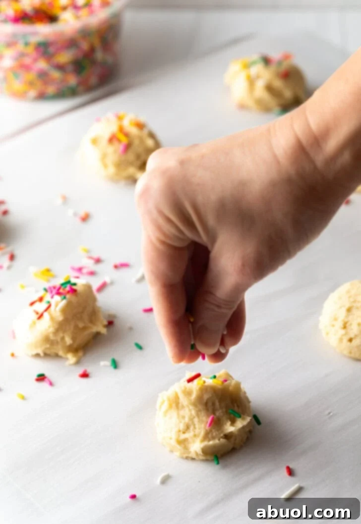 Luscious Cream Cheese Delights 6 Hand adding colorful sprinkles to the tops of raw cookies balls spread out on a baking sheet.