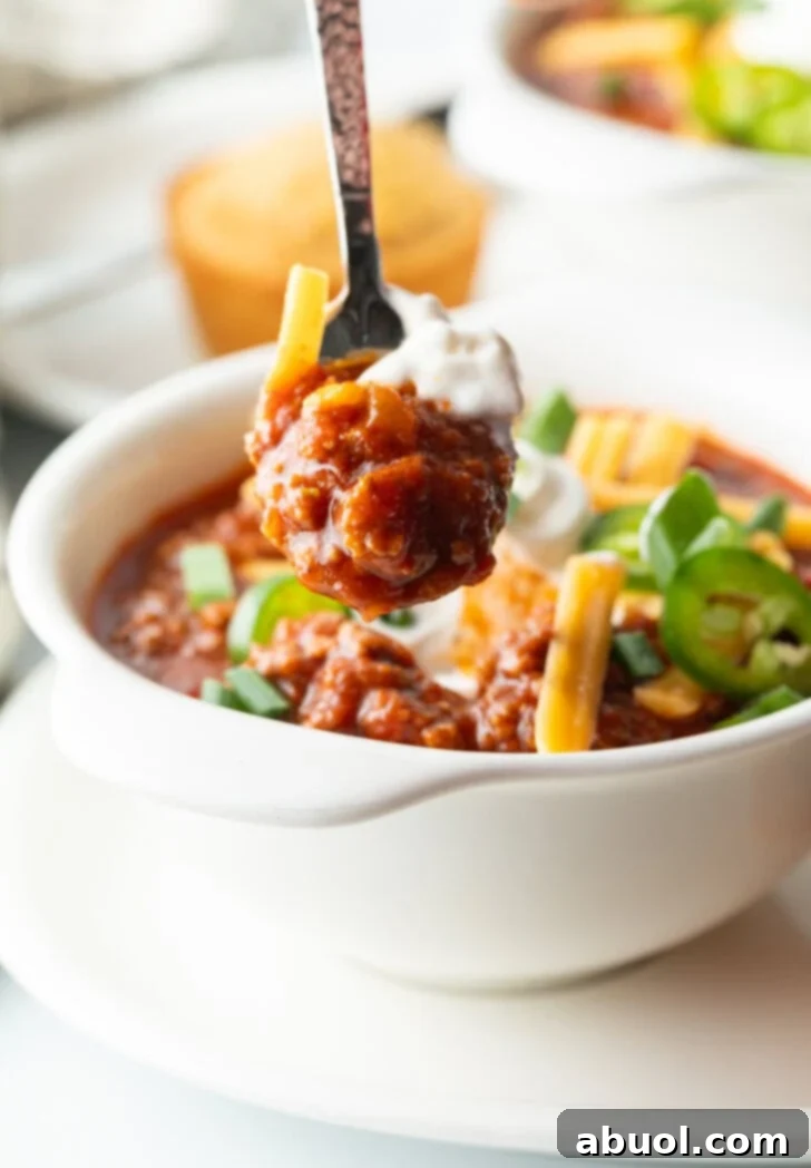 White bowl of chili no bean chili topped with shredded cheese, jalapeno slices, sour cream and chopped green onions. A spoon is showing a spoonful of chili con carne to camera.