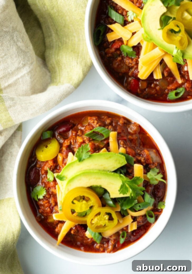 Top down bowl of venison chili with pickled jalapeno peppers, shredded cheese, slices of avocado and chopped green onions