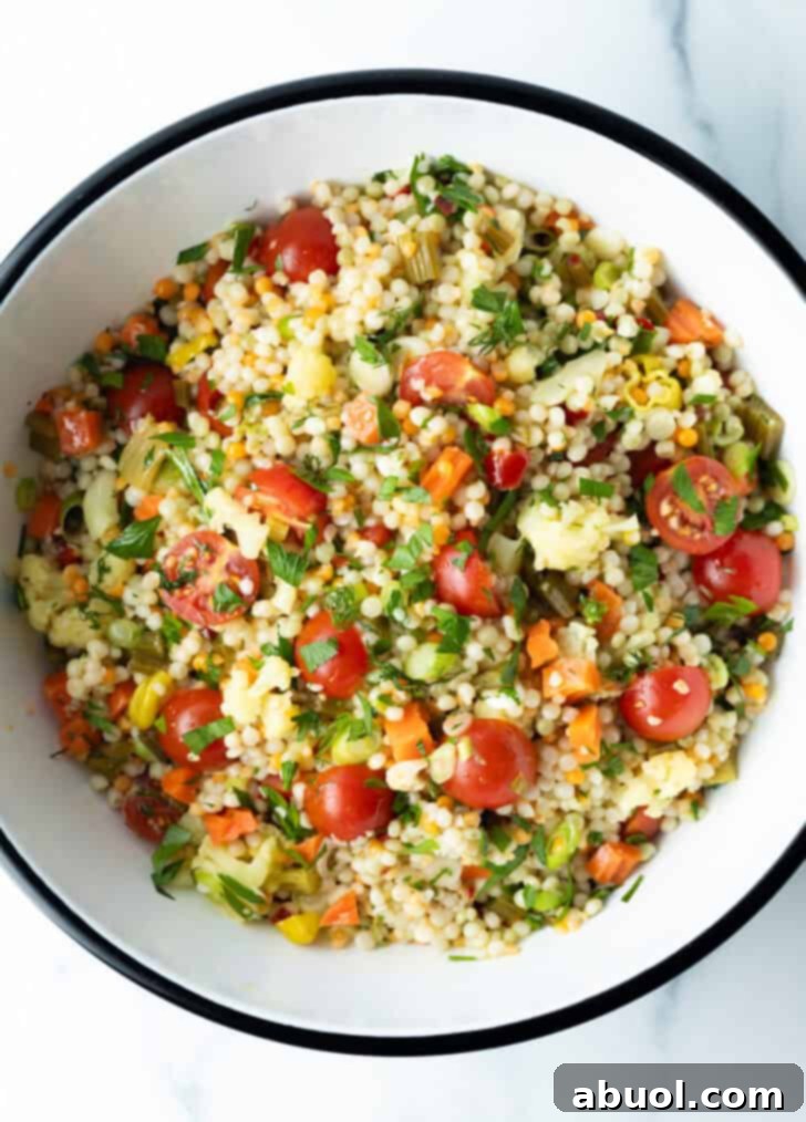 Top-down view of a colorful pearl couscous salad in a large white mixing bowl, featuring chopped tomatoes, fresh herbs, and tangy pickled vegetables.