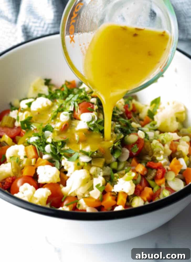 Pouring the vibrant, homemade dressing over a colorful mixture of chopped vegetables and pearl couscous in a large mixing bowl, ready to be tossed.