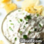 Bowl of creamy white French onion dip, topped with chopped chives and with a potato chip stuck into the dip.