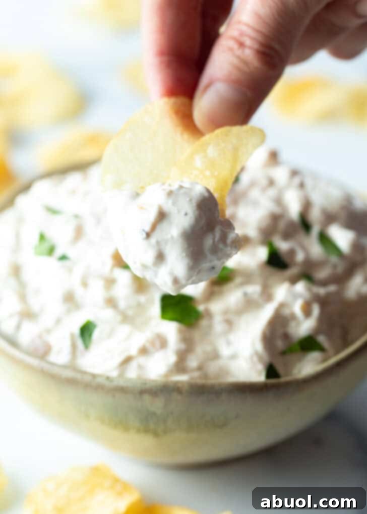 A hand holding a potato chip scoops a generous dollop of homemade French onion dip towards the camera, showcasing its creamy texture.