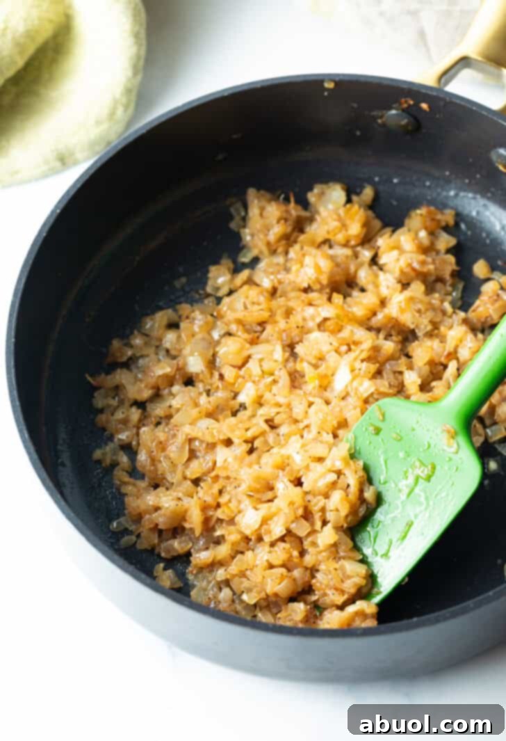 Caramelizing finely diced onions in a large skillet with a green spatula, showing them beginning to soften and brown.