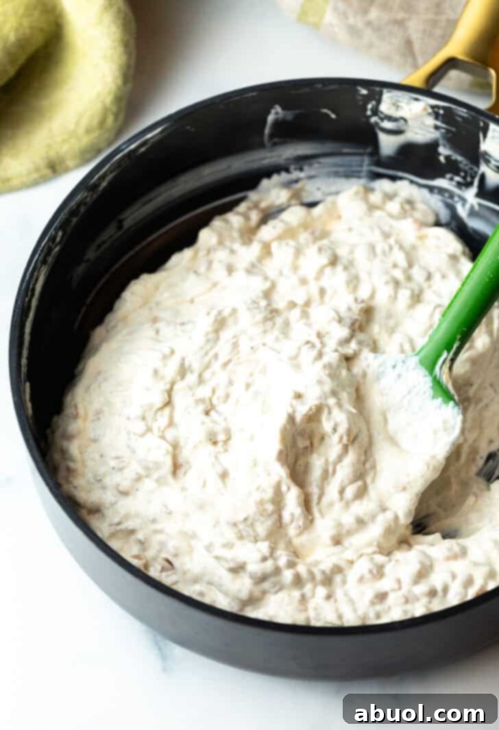 Stirring the creamy French onion dip in a large pot with a green spatula, showing the smooth, well-combined texture.