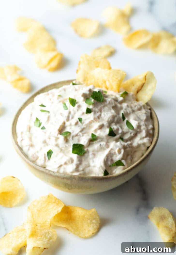 Bowl of creamy white French onion dip, perfectly garnished with chopped chives and featuring a crisp potato chip enticingly dipped into the creamy mixture.