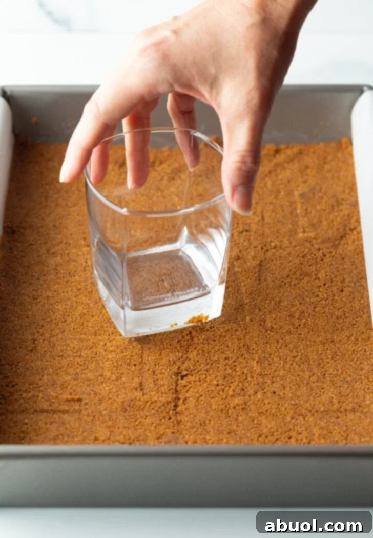 A hand using a drinking glass to press the gingersnap cookie crust into a baking dish.