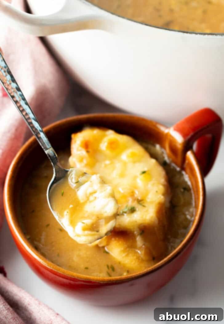 A large red mug of French onion soup with a cheese-topped baguette, a spoon dipping in for a delicious scoop.