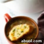 A comforting large red mug of French onion soup, elegantly topped with a toasted baguette slice and melted Gruyere cheese.