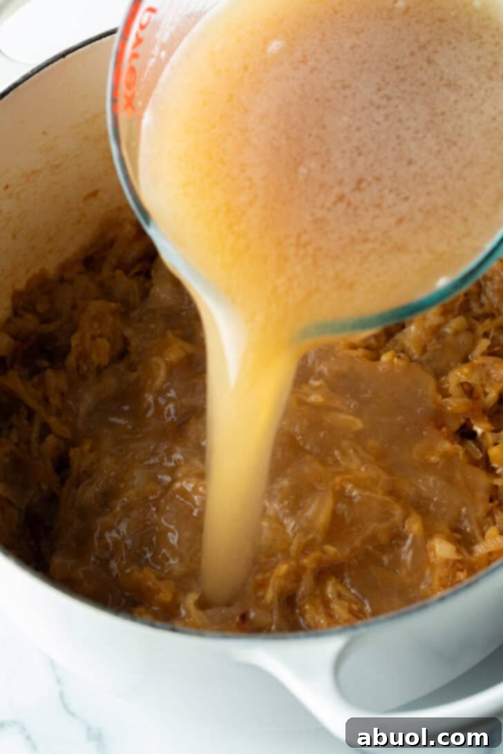 A glass pitcher carefully pouring rich beef stock into the large pot, where the cooked, tender onions await, ready to create the flavorful soup base.