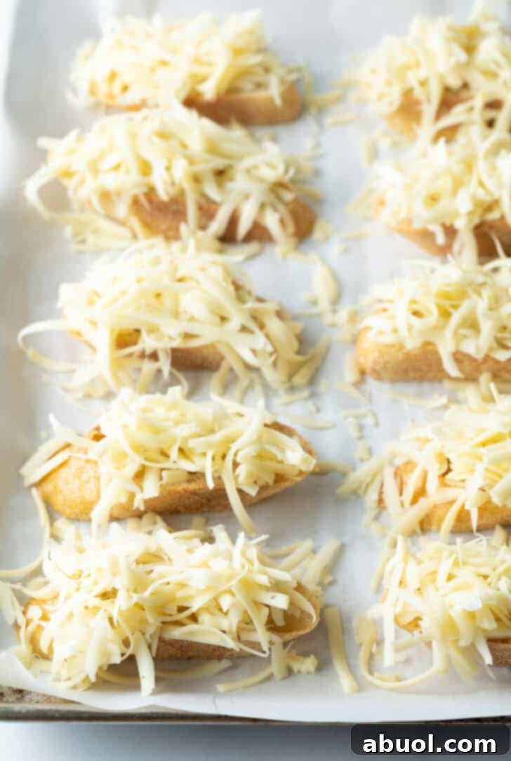 A close-up of eight perfectly sliced baguette pieces arranged on a baking sheet, each generously topped with a mound of shredded cheese, ready for broiling.