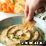 Hand using a pita chip to scoop eggplant dip from a light blue bowl.