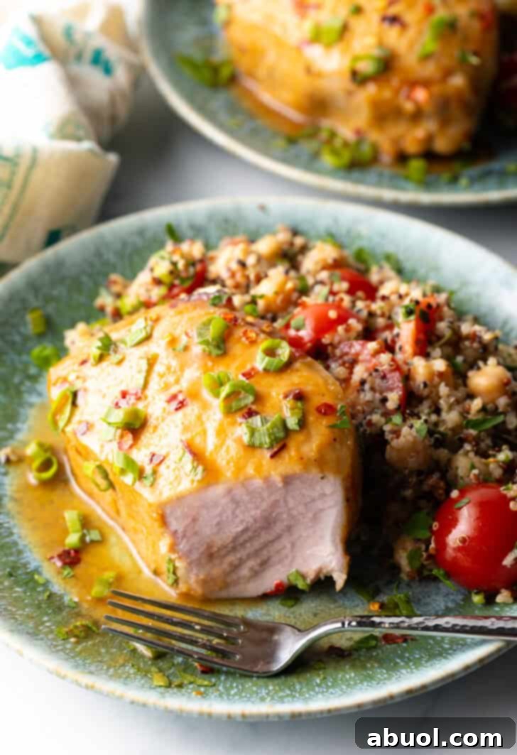 Perfectly Baked Pork Chops 11 A close-up view of a baked pork chop, expertly glazed and garnished with fresh scallions, on a plate next to a vibrant quinoa salad. A portion of the chop has been cut to reveal its succulent, perfectly cooked interior.
