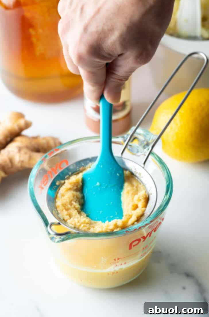 A blue spatula actively pushing pureed ginger mixture through a fine-mesh sieve into a bowl, separating pulp from juice.