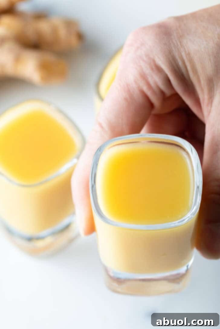 A hand holding a shot glass filled with golden yellow ginger shot liquid, offering it towards the camera.