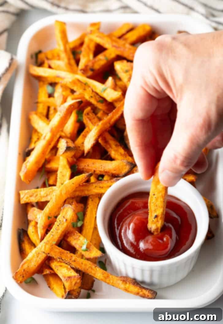 Hand dipping a sweet potato fry from air fryer in a white ramekin of ketchup.