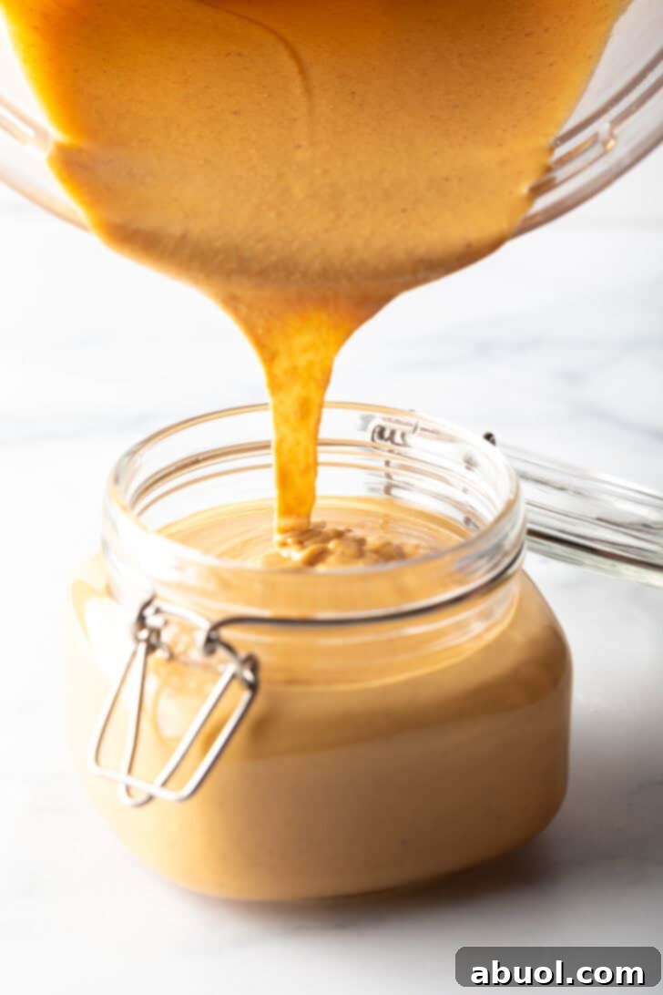 Pouring smooth homemade peanut butter from a blender jar into a glass storage jar, demonstrating its perfect pourable consistency.