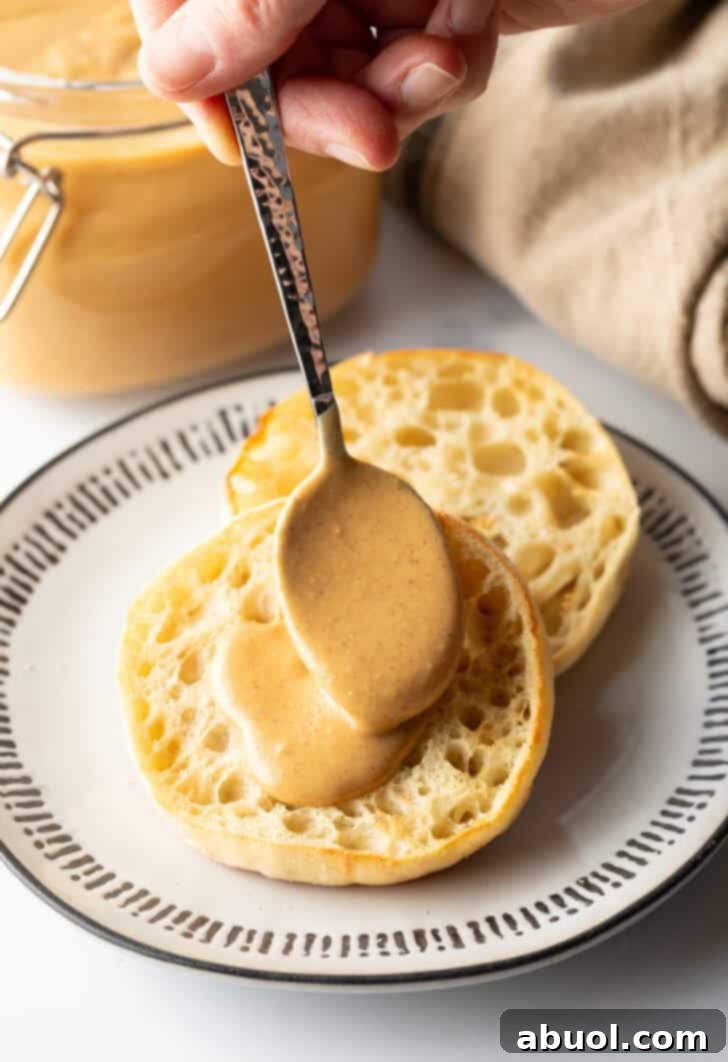 A hand using a spoon to smooth freshly made homemade nut butter onto half of an English muffin, ready to be enjoyed.