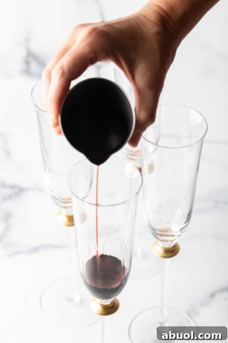 Hand pouring deep red liquid from a metal measuring cup into a fluted champagne glass.
