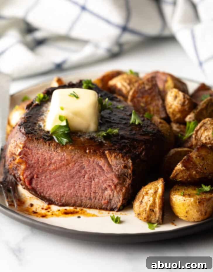 White plate with a reverse sear steak, cut in half to show the medium-rare meat inside. The steak is topped with chopped herbs and a pat of butter. Roasted yellow potato pieces are on the right side of the meat.
