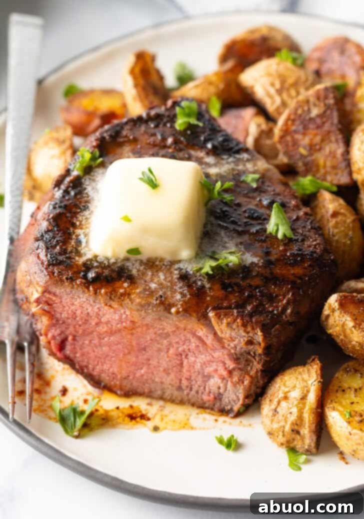 White plate with a reverse sear steak, cut in half to show the medium-rare meat inside. The steak is topped with chopped herbs and a pat of butter. Roasted yellow potato pieces are on the right side of the meat.
