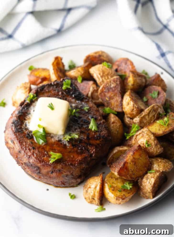 White plate with a reverse sear steak, topped with chopped herbs and a pat of butter. Roasted yellow potato pieces are on the right side of the meat.