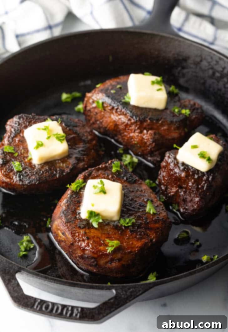 Four filets searing in a cast iron skillet. Each steak is topped with fresh chopped herbs and a pat of butter.
