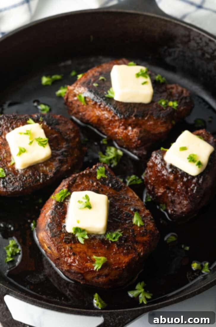 Four filets searing in a cast iron skillet. Each steak is topped with fresh chopped herbs and a pat of butter.