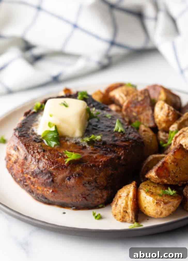 White plate with a reverse sear steak, topped with chopped herbs and a pat of butter. Roasted yellow potato pieces are on the right side of the meat.