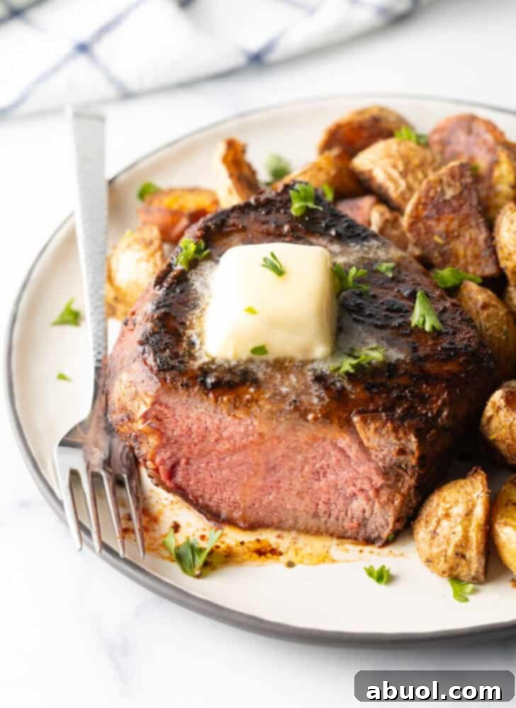 White plate with a reverse sear steak, cut in half to show the medium-rare meat inside. The steak is topped with chopped herbs and a pat of butter. Roasted yellow potato pieces are on the right side of the meat.