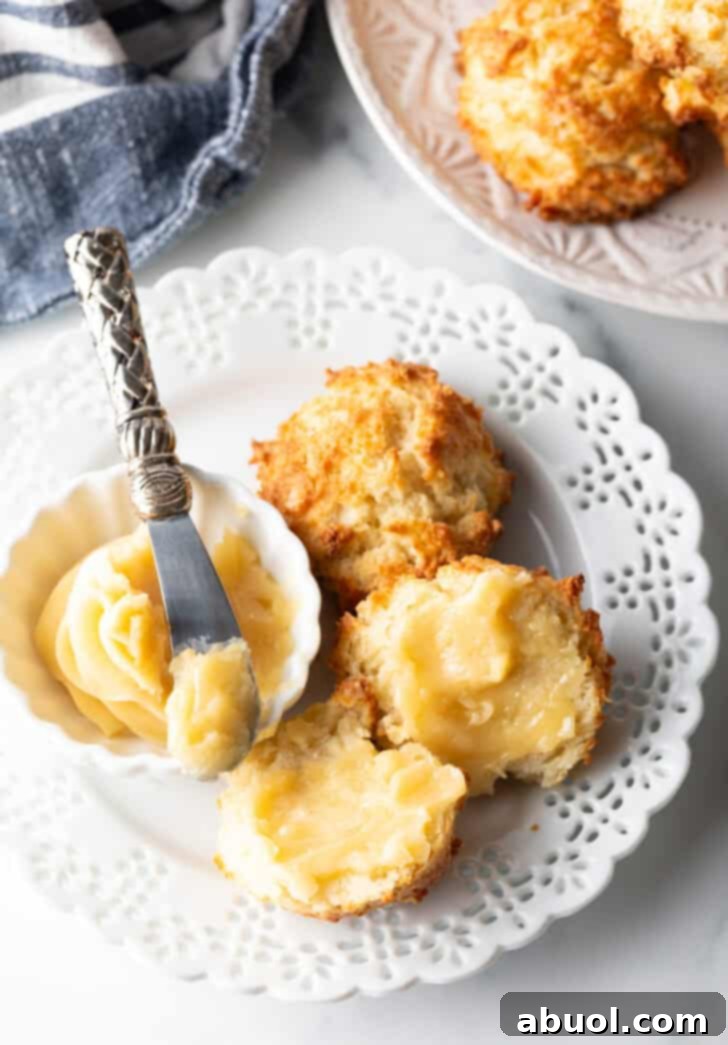 Close view of two biscuits on a white plate, with a white ramekin of whipped honey butter and a butter knife showing a bit to camera. One biscuit is open with both sides buttered.