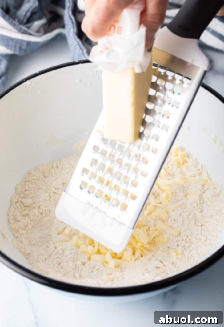 Using a cheese grater to grate butter into a white bowl with dry biscuit ingredients.