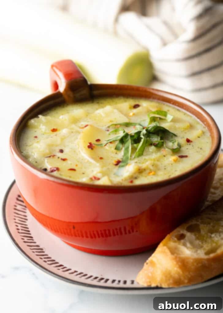 Red oversized mug filled with potato leek soup, on a plate with slice of baguette on the side.