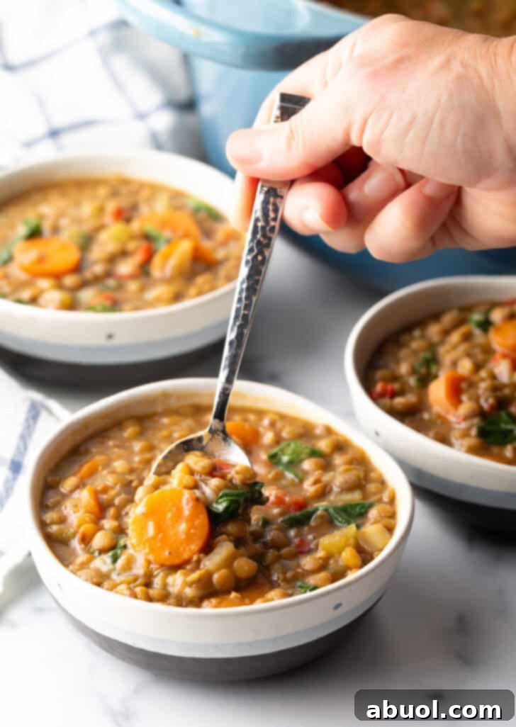 A hand holding a spoon, scooping a generous serving of traditional lentil soup from a white bowl, ready to be enjoyed.