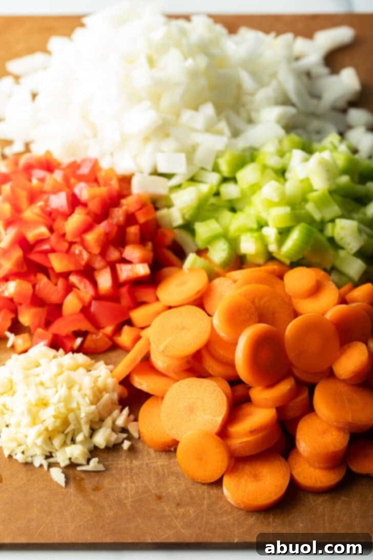 Chopped fresh ingredients in neat piles on a wood cutting board: Onion, red bell peppers, carrots, minced garlic, and sliced celery, perfectly prepped for cooking.